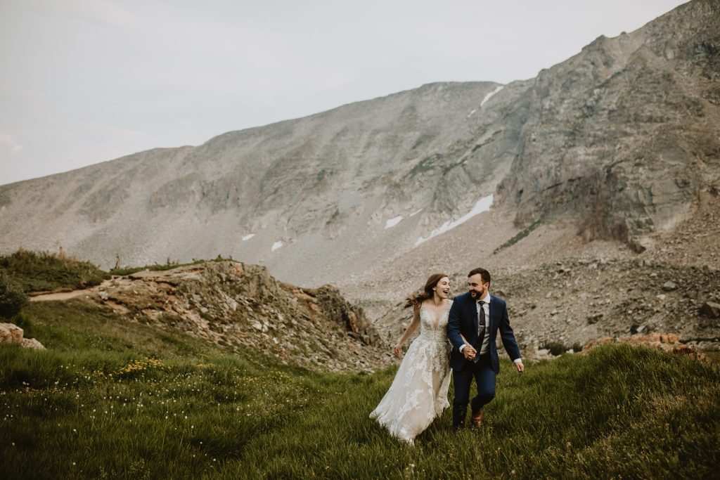 Couple in wedding attire running through mountain grasses during wedding ceremony
