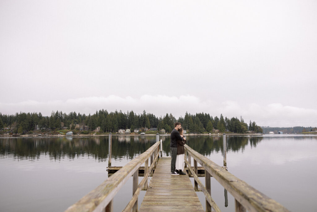 A couple stands on a long wooden dock, embracing, looking into distance with a calm lake and a forested shoreline in the background.
