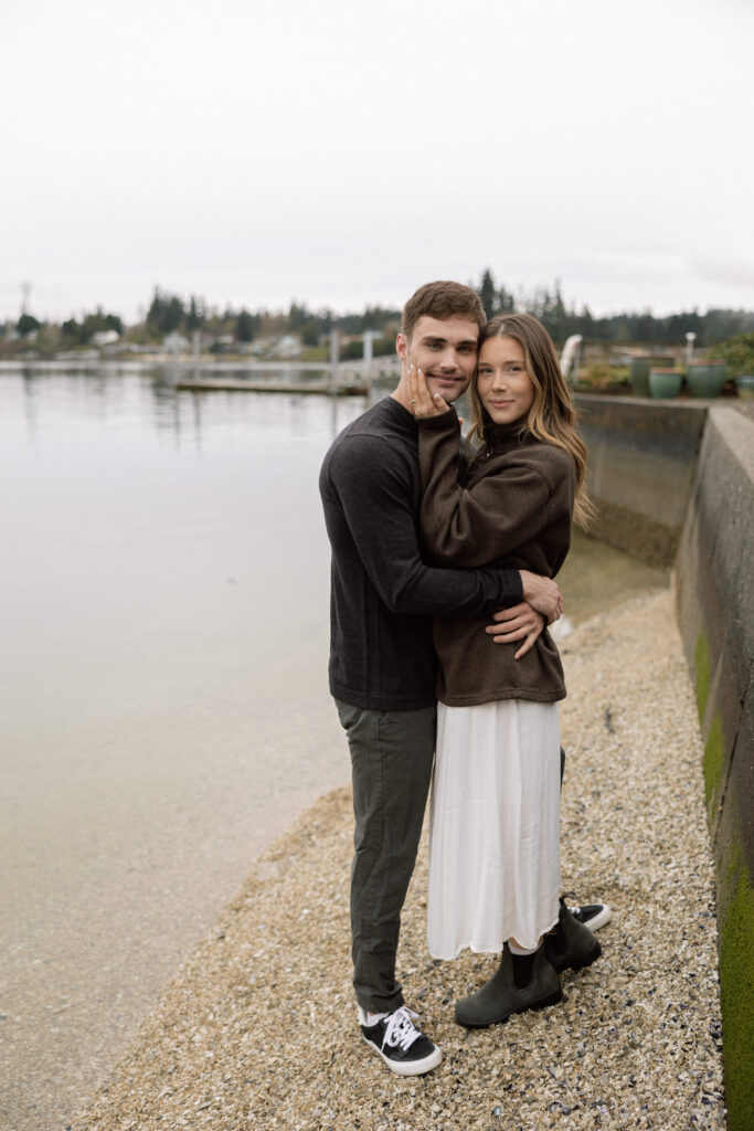 A couple stands by the shoreline, with the woman gently touching the man's face as they both look at the camera. They are dressed in casual, comfortable clothing, and the calm water and dock in the background add to the serene atmosphere.