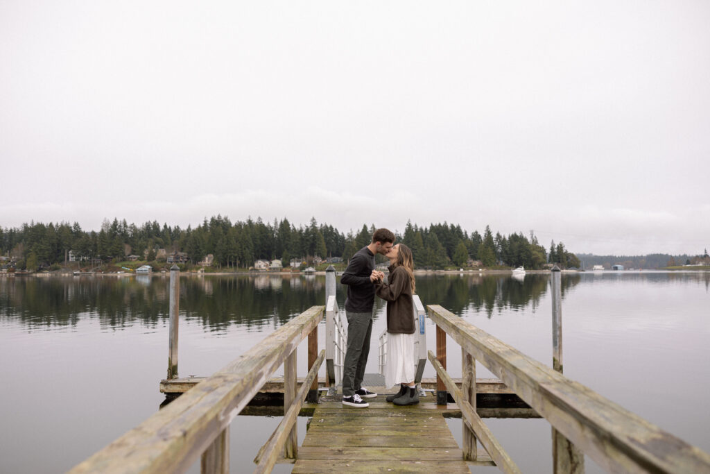 A couple stands at the end of a wooden dock, sharing an intimate moment by a tranquil lake with forested shores and houses in the background.