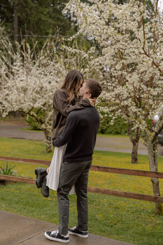 A couple shares a kiss under blooming trees, with the man lifting the woman slightly off the ground. The background features white blossoms and greenery, creating a romantic and picturesque scene