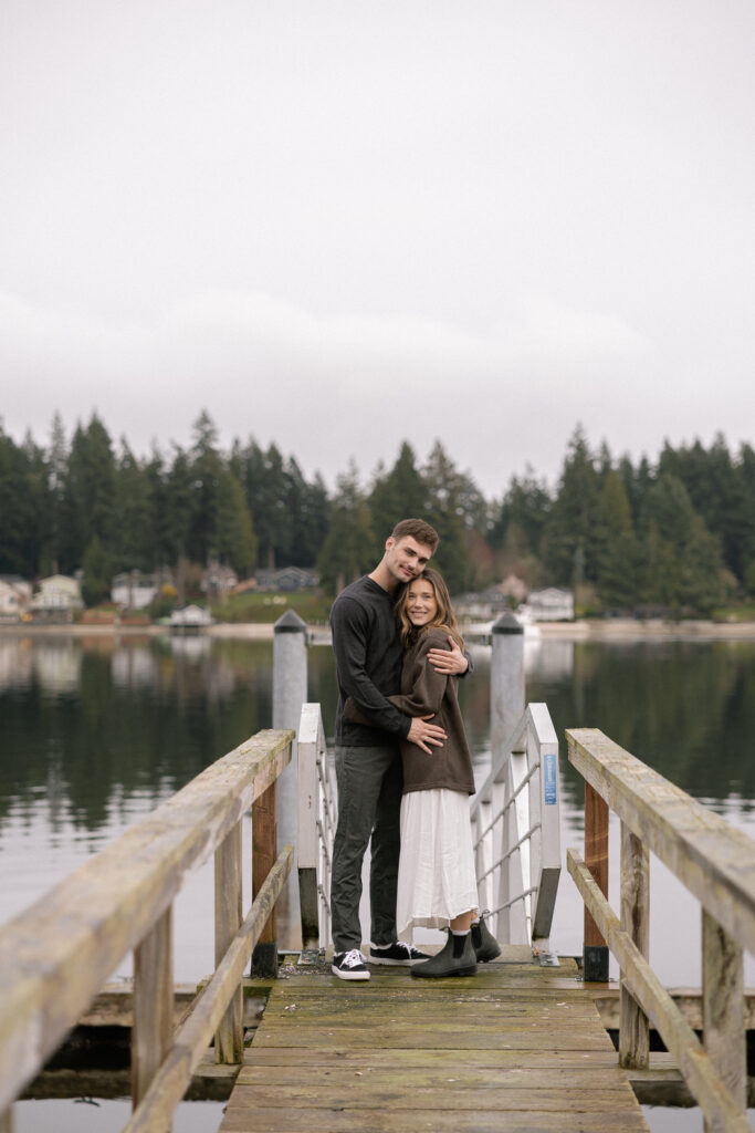 A couple stands on a wooden dock, embracing and smiling, with a serene lake and forested background.